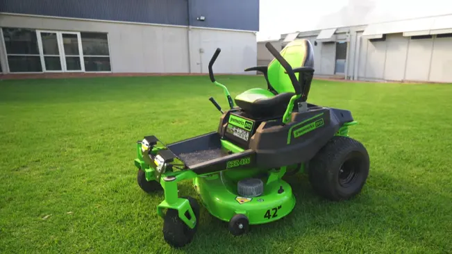 green riding lawnmower with black seating and handles, parked on a lush, green lawn in front of a grey building with large windows.