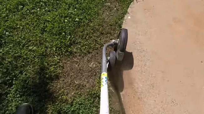 First-person perspective of hands holding a lawnmower handle, ready to mow overgrown grass beside a dirt path