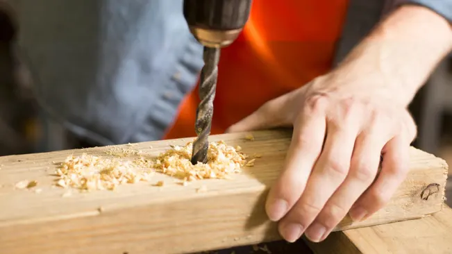 Person using a drill on a wooden plank, creating wood shavings