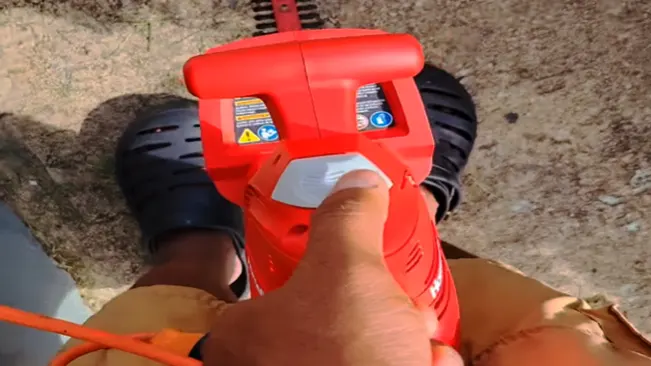 Hand operating a hedge trimmer safety labels, wearing black shoes and brown pants