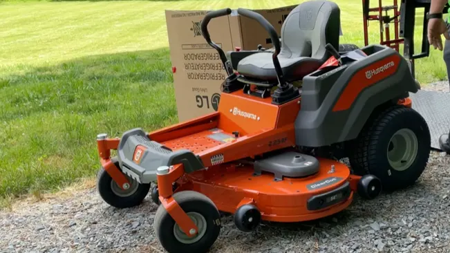 brand-new Husqvarna riding lawnmower, predominantly orange with black accents, parked on a gravel surface with a lush green lawn