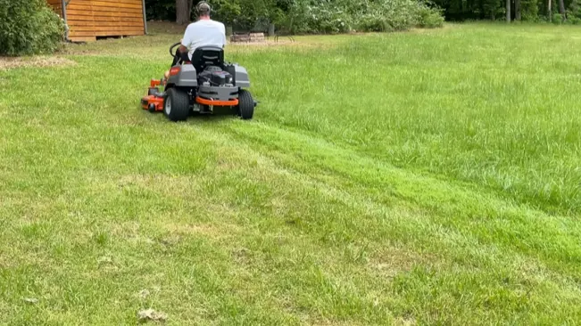 person on an orange and black riding lawnmower, cutting grass in a large yard with trees and a wooden structure