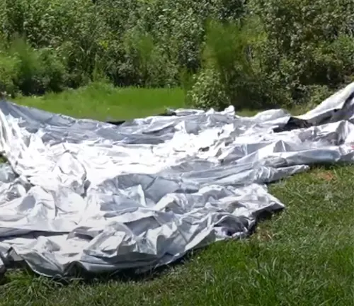 A deflated paint booth lies on the grass, its silver material spread out and crinkled, awaiting inflation, set against a backdrop of lush greenery.