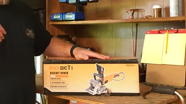 Person holding a REX BETI Biscuit Joiner box near a wooden shelf and toolbox