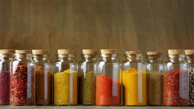 A close-up of a row of glass bottles filled with powdered spices on a wooden table.
