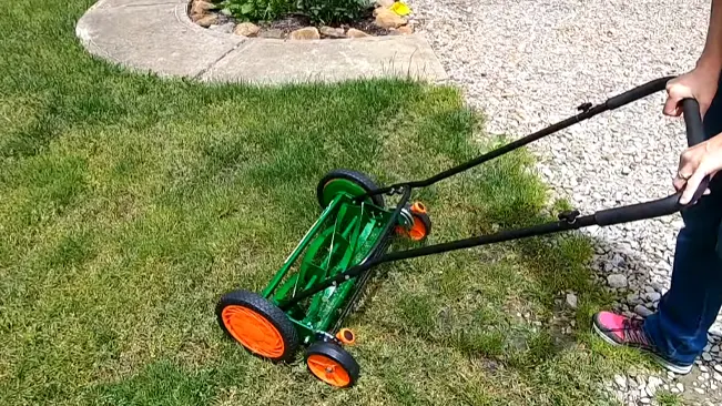 visible from the waist down, pushing a green manual lawn mower with bright orange wheels across a well-maintained lawn on a sunny day