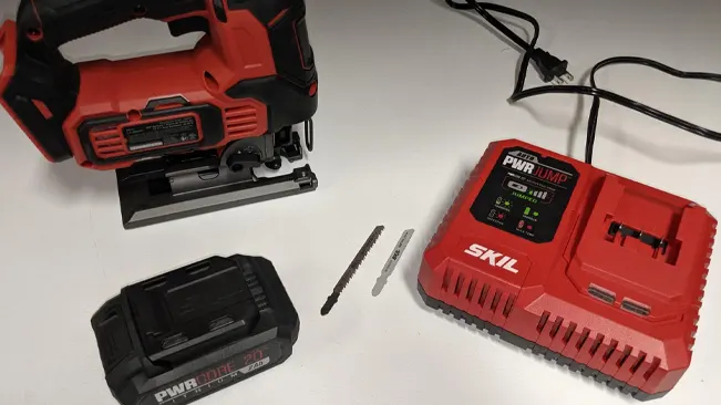 Red and black jigsaw, two jigsaw blades, a battery, and a battery charger on a white surface