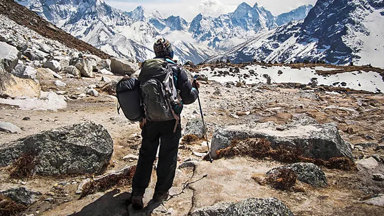 A solitary hiker with a large backpack and trekking poles standing on a rocky path, gazing out at a rugged, snowy mountain landscape.
