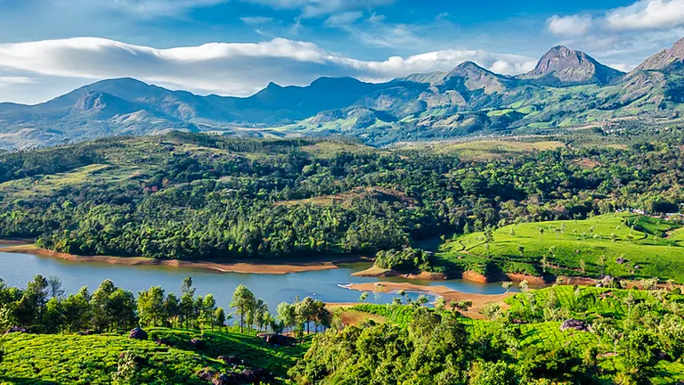 Vibrant landscape showcasing the lush Western Ghats with rolling hills covered in tea plantations, a serene lake in the foreground, and rugged mountain peaks in the backdrop under a clear sky.
