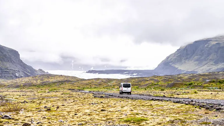 A vehicle on a remote gravel path with a backdrop of misty mountains and a glacier in the distance.
