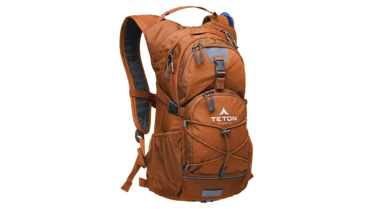 A burnt orange TETON sports hiking backpack with a blue hydration bladder tube, featuring multiple zippered compartments, adjustable straps, and a bungee cord attachment system on the front. The pack has a mesh back panel for breathability and side pockets for additional storage.