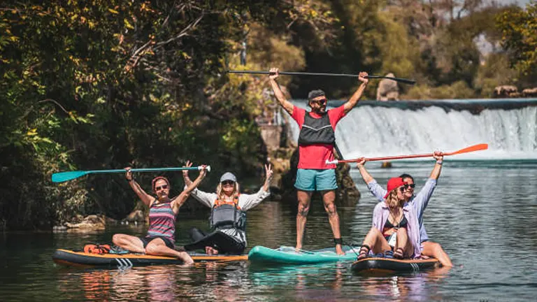 Four people joyfully paddling on colorful stand-up paddleboards and kayaks on a river, with lush greenery in the background and a waterfall in the distance.