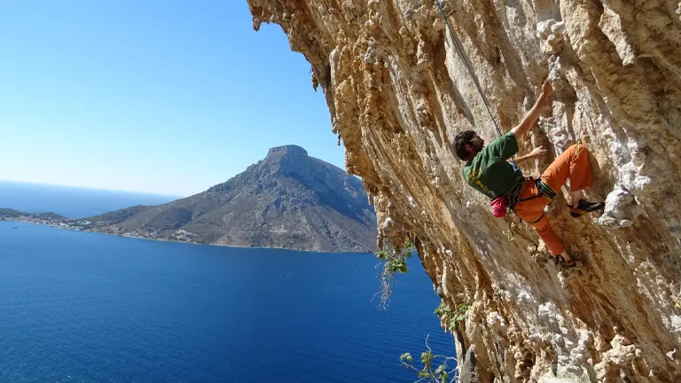A climber on an overhanging, rugged rock face with a scenic ocean backdrop, under a clear sky.

