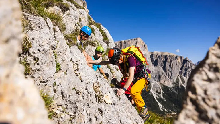 Two climbers in colorful gear ascending a rocky mountain slope with a steep cliff in the background under a clear blue sky.


