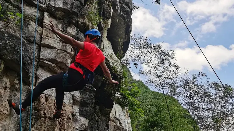A climber in a red shirt and blue helmet ascends a rock face with lush greenery in the background.

