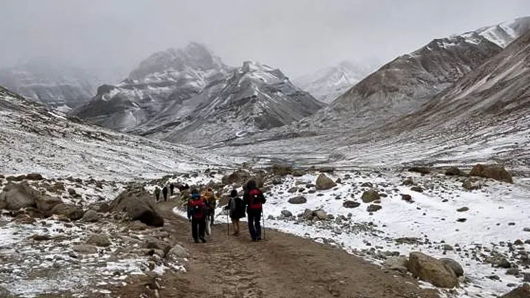 A group of hikers trekking on a rocky path through a snow-dusted valley with rugged mountains shrouded in mist in the background.
