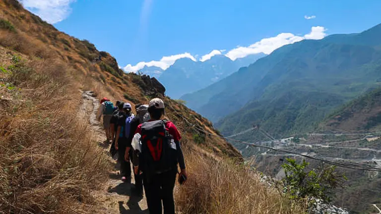 Hikers in a single file trekking on a narrow dirt trail along a grassy hillside with panoramic views of deep valleys and distant mountains under a clear sky.


