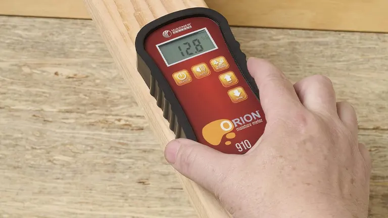 A person's hand pressing buttons on a red and black Orion 910 pinless moisture meter held against a piece of lumber, with a moisture level of 12.8% shown on the digital display.
