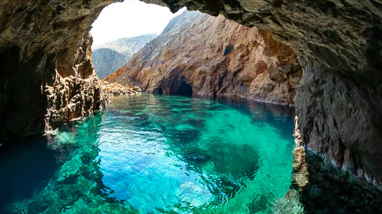 A view from inside a sea cave looking out to a turquoise cove surrounded by rocky cliffs, illuminated by natural light.
