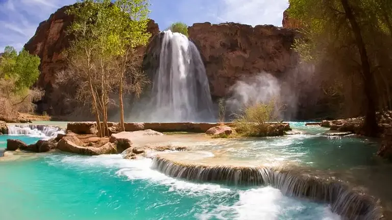 A breathtaking view of Havasu Falls with its turquoise waters cascading down red rock cliffs, surrounded by green vegetation under a clear blue sky.


