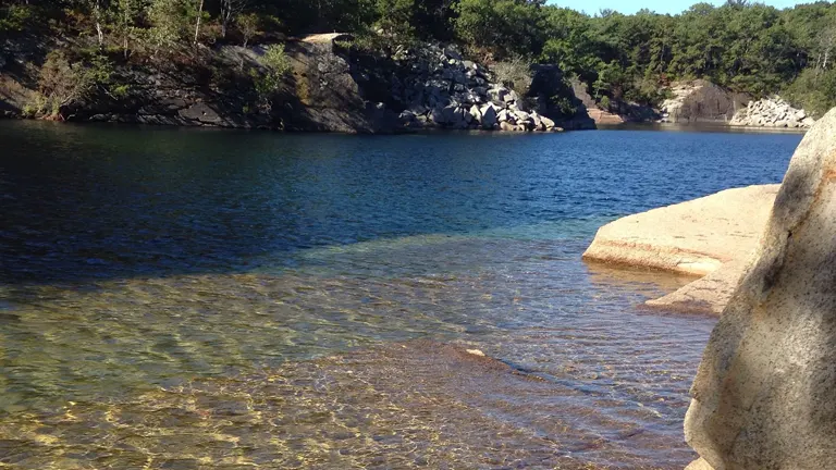 A clear and shallow entry to a serene quarry lake with deep blue water, surrounded by rocky outcrops and pine trees under a bright blue sky.


