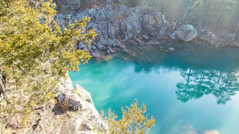A serene view of a clear, turquoise mountain lake with reflections of surrounding pine trees on the water's surface. Sunlight filters through the forest, illuminating the rocky terrain and evergreens at the water's edge.