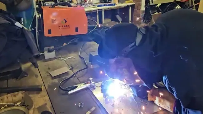 A person in protective gear welding with a HITBOX MIG200II MIG welder, with bright sparks flying, in a workshop environment.