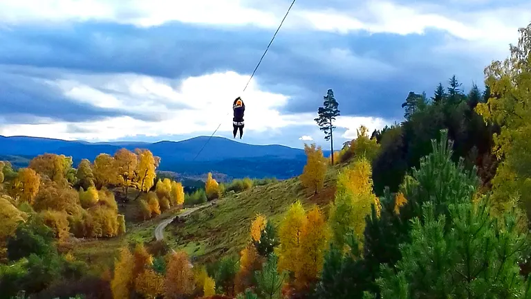 A person is zip-lining over a scenic landscape adorned with autumn-colored trees, rolling hills, and a cloudy sky above.
