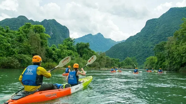 A group of kayakers wearing helmets and life jackets paddles down a gentle river, flanked by lush greenery and mountainous terrain.

