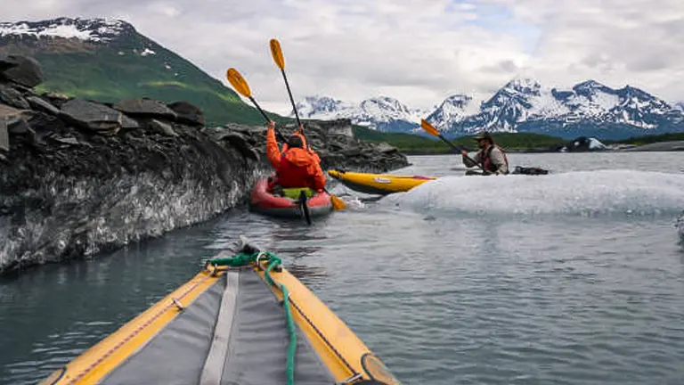 Kayakers navigating icy waters near glaciers with snow-capped mountains in the background, indicative of a cold, alpine environment.

