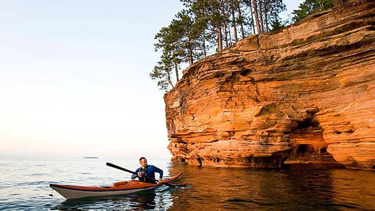 A kayaker paddles along a calm lake next to a towering sandstone cliff with trees at the top during a peaceful evening.
