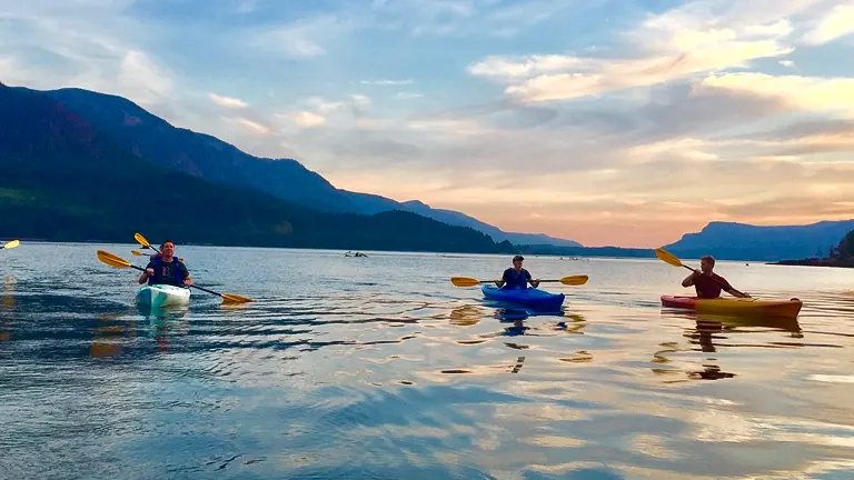 A group of kayakers on a serene lake at dusk, with the water reflecting the pastel hues of the sunset and mountains silhouetted against the sky.

