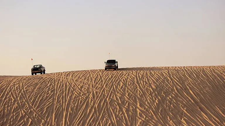 Two vehicles with safety flags cresting a rippled sand dune under a clear sky, highlighting the vastness and isolation of the desert environment.

