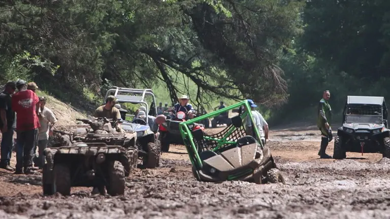 A group of people gathered around off-road vehicles on a muddy trail, with one vehicle tilted to its side, suggesting a rollover incident while navigating the challenging terrain.

