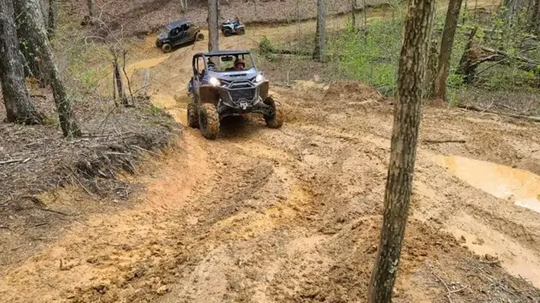 A UTV tackling a steep, sandy incline in a forested off-road area, with other vehicles waiting their turn on the trail below.

