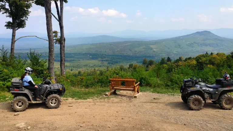 A rider on an ATV pauses on a scenic overlook with expansive views of rolling hills and a clear sky, with another ATV parked beside a wooden bench.
