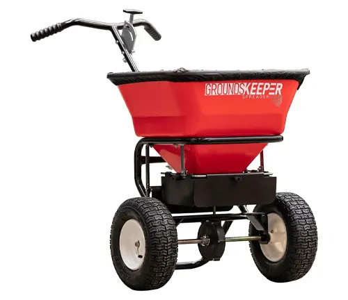 A red Groundskeeper broadcast spreader with a large hopper and pneumatic tires, featuring a black frame and handle.
