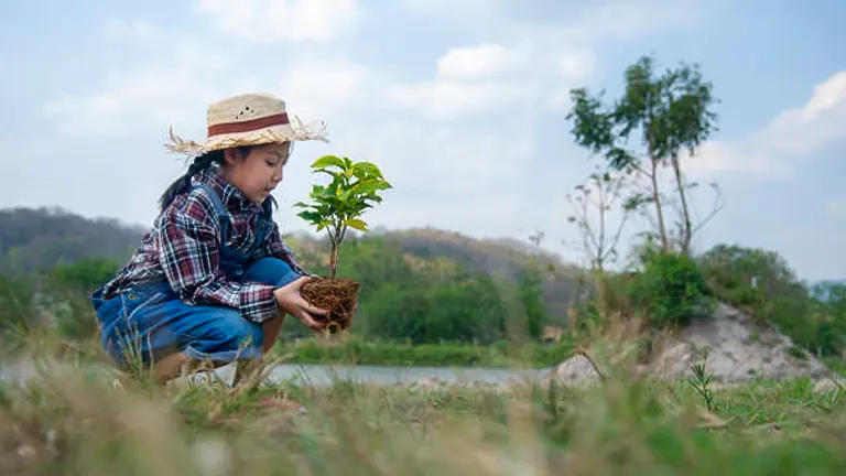 A child in a straw hat kneeling to plant a young tree outdoors, with a scenic backdrop of a clear sky and rolling hills.