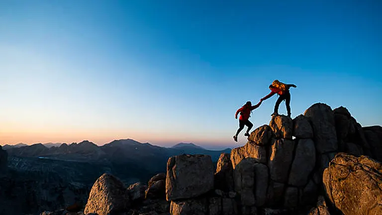 Two climbers on a rocky summit during sunset, with one reaching down to assist the other against a backdrop of a mountainous horizon.
