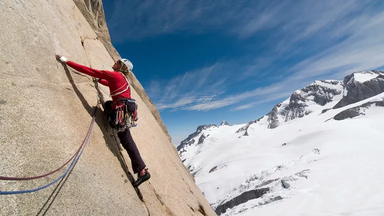 A climber in a red top and black pants is scaling a steep granite rock face with a backdrop of snow-covered mountains and a clear blue sky.
