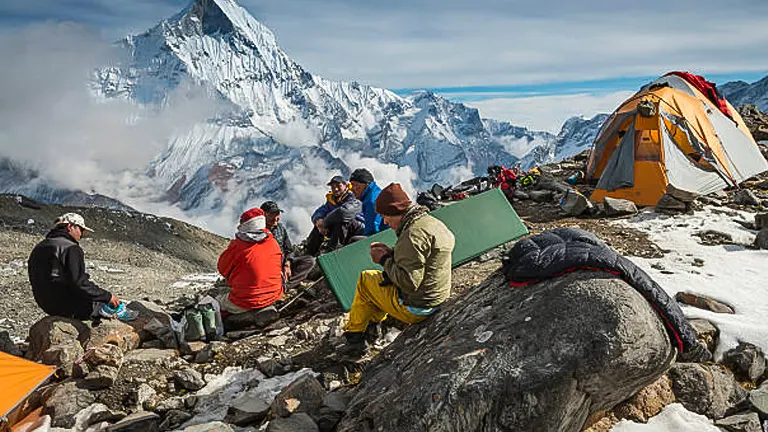 A group of hikers resting at a high-altitude campsite with snow-covered peaks in the background, tents pitched on rocky terrain, and clouds swirling below them.
