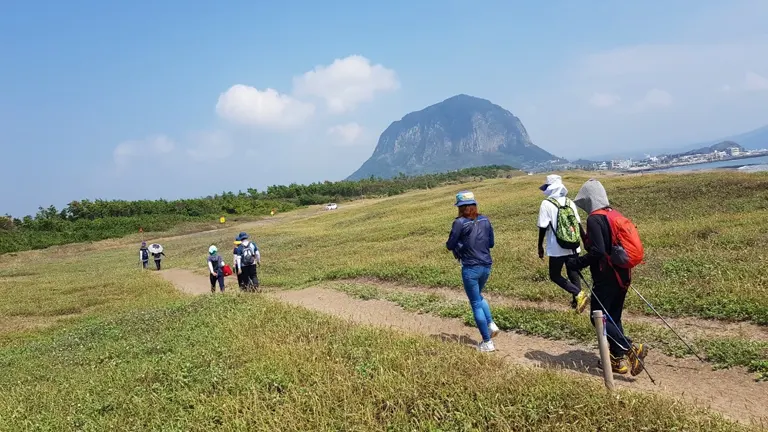 A group of casual hikers walking on a grassy trail leading towards a distinctive, solitary mountain under a clear blue sky.

