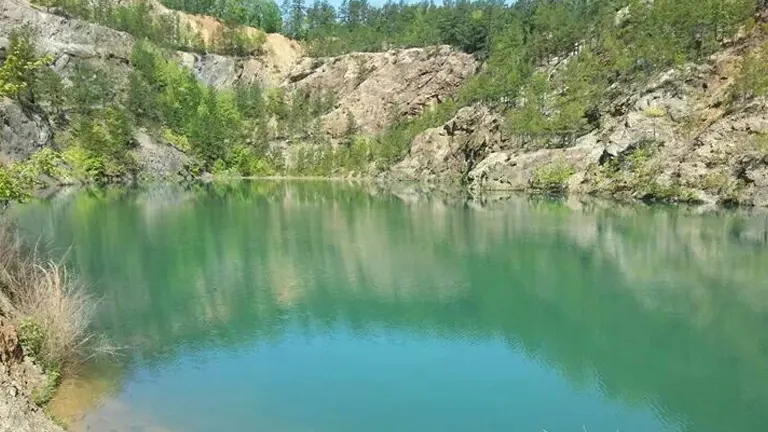 A quarry lake with emerald-green waters nestled within a rocky landscape, under a clear blue sky, presenting a serene and inviting natural setting.

