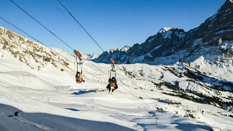 People ziplining over a snowy mountain landscape with alpine peaks in the distance and a clear blue sky.