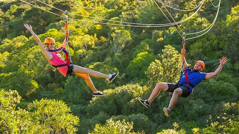 Two people ziplining side by side, smiling and posing with their arms outstretched, over a lush green forest.