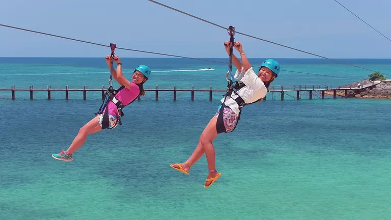 Two people, each wearing helmets and harnesses, are enjoying a zip-line adventure above crystal clear turquoise waters near a coastal area.
