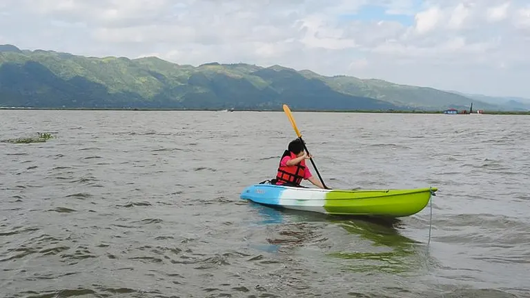 A person in a life jacket paddles a bright green kayak on a vast lake with rolling hills in the background under a cloudy sky.

