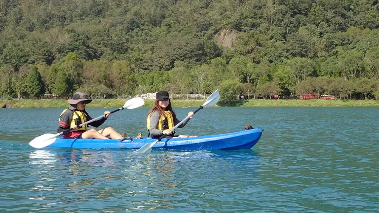Two people in a blue tandem kayak paddle on a calm, clear lake with lush greenery and hills in the background on a sunny day.

