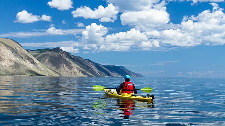 A lone kayaker in a red life jacket on a yellow kayak glides across still waters with mountainous terrain in the background under a vast sky dotted with clouds.
