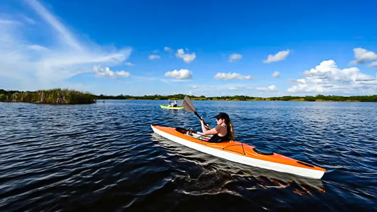 Two people in kayaks are paddling on a wide, sunlit river with lush greenery on the distant banks under a blue sky with wispy clouds.
